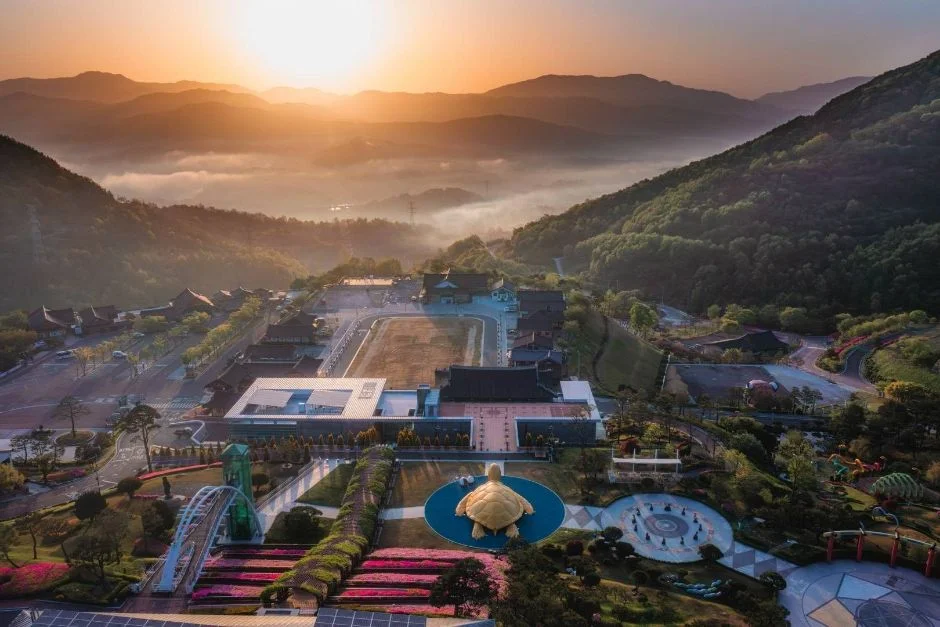 일출 무렵 안개 낀 산자락 아래 건물과 황금 거북 조형물이 보이는 동의보감촌 항공 사진