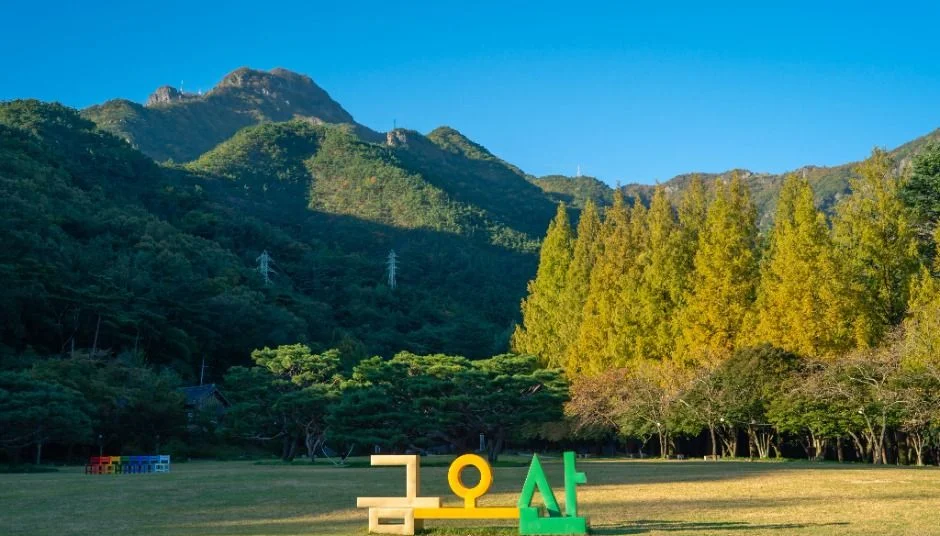 금오산 글자 조형물과 푸른 산, 노란 나무가 보이는 잔디밭