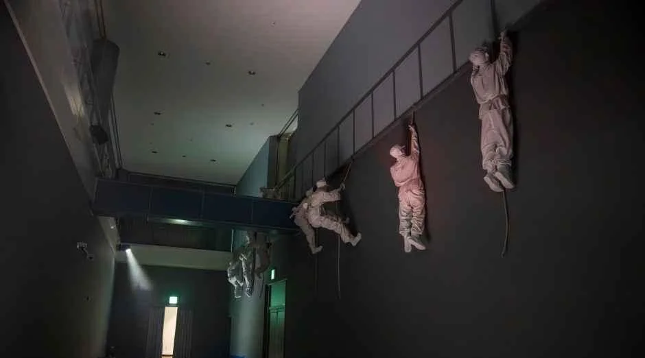 어두운 전시 공간 벽면을 오르는 흰색 인물 조형