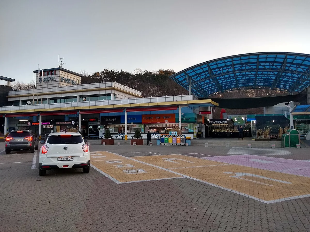 고속도로 휴게소 매점 건물과 파란 지붕, 앞쪽에 승용차가 서 있는 주차 구역