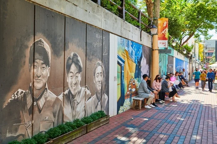 여러 인물 초상이 그려진 벽 앞 벤치에 사람들이 앉아 있는 골목