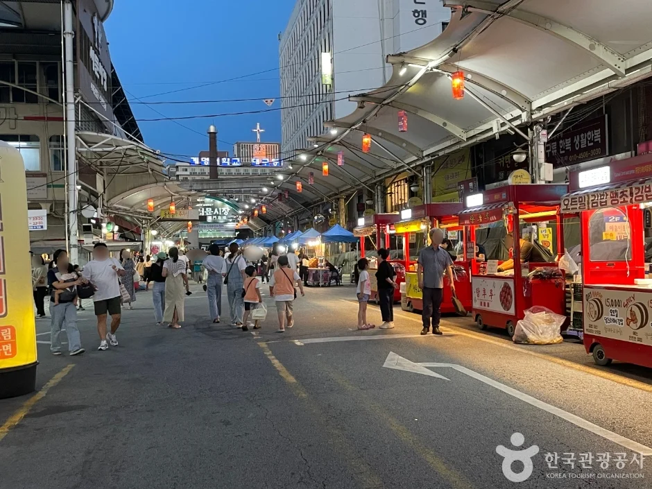 어둑한 저녁 하늘 아래 아케이드 거리에 사람들이 모여 걷는 야시장 풍경