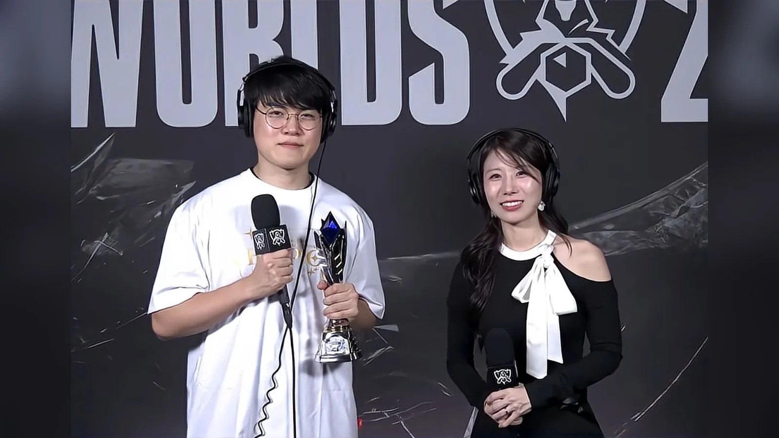 월드 챔피언십 로고가 보이는 배경 앞에서 흰 티셔츠 차림의 남성이 트로피와 마이크를 들고 여성 진행자 옆에 서 있는 인터뷰 장면
