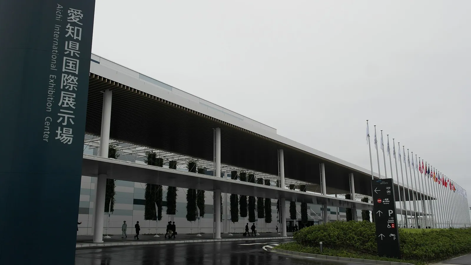 愛知県国際展示場 안내 간판이 선 진입로 너머로 처마가 긴 전시장 외관과 줄지어 선 깃대가 보이는 모습