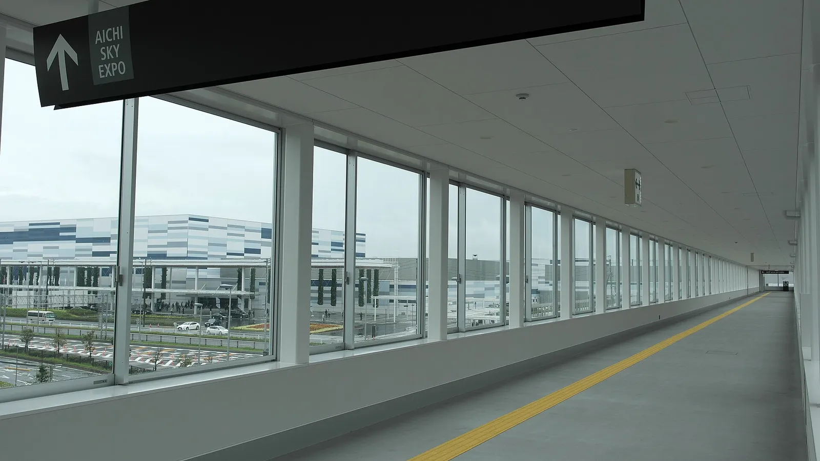 AICHI SKY EXPO 안내 표지가 달린 유리창 연결 통로 너머로 푸른 줄무늬 외벽의 전시장이 보이는 모습