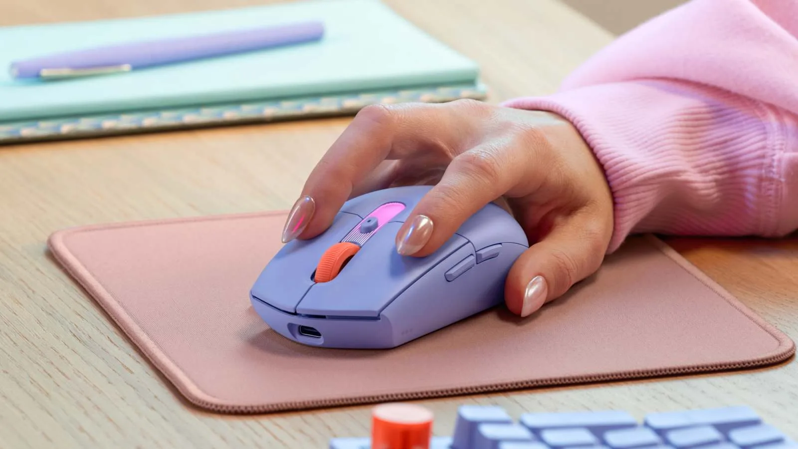 로지텍 G304X 수퍼라이트 라일락 무선 마우스를 사용자가 손으로 쥐고 마우스패드 위에서 사용하는 모습, 책상 위 파스텔톤 노트와 펜이 함께 놓여 있다