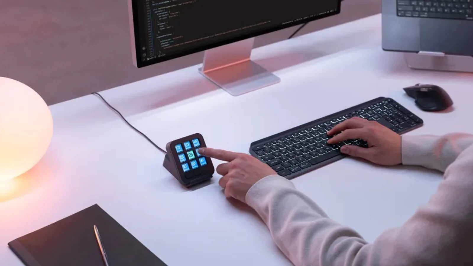 로지텍 MX Keypad 실사용 이미지 - 책상 위에서 사용자가 검지로 그래파이트 색상 키패드의 풀컬러 LCD 키를 누르는 모습, 뒤로 코드 에디터가 켜진 모니터