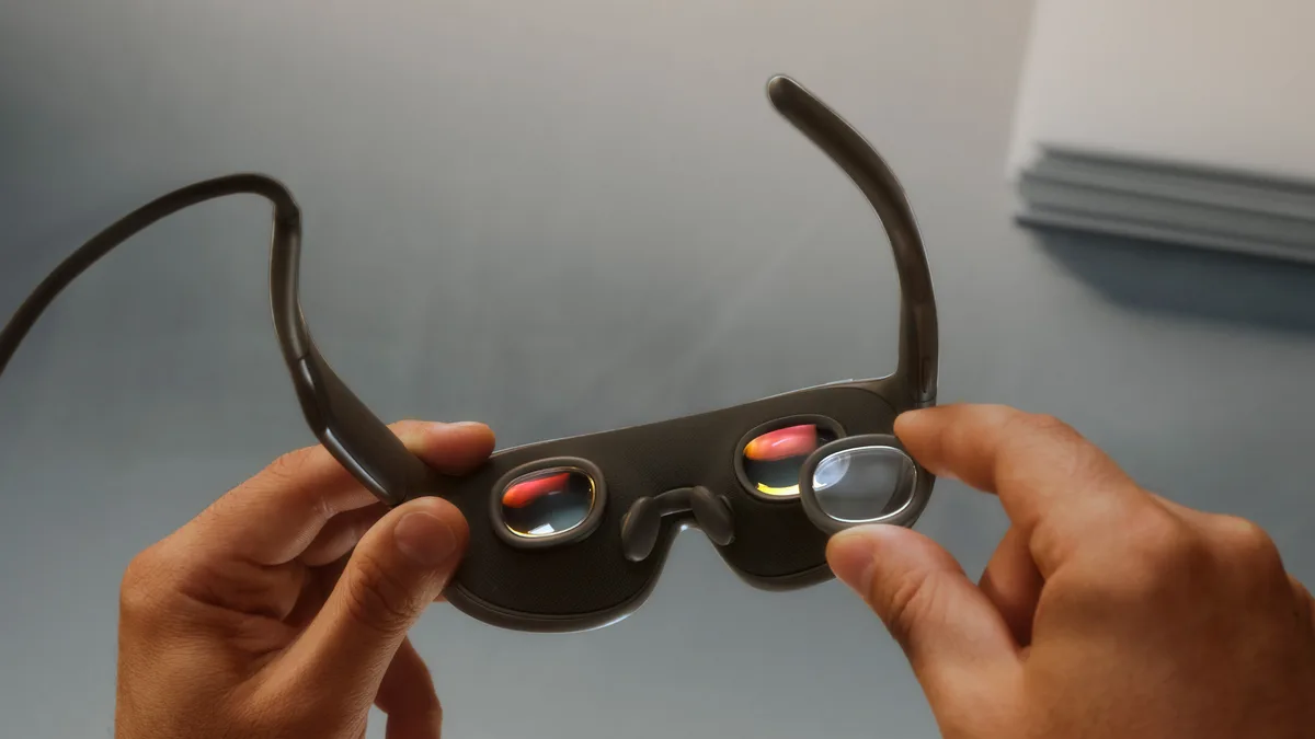 손으로 메타 VR 글래스 렌즈 부분을 살펴보는 모습 - 안쪽 렌즈 두 개가 보이고 책상 위에는 별도의 일반 안경이 놓여 있는 모습