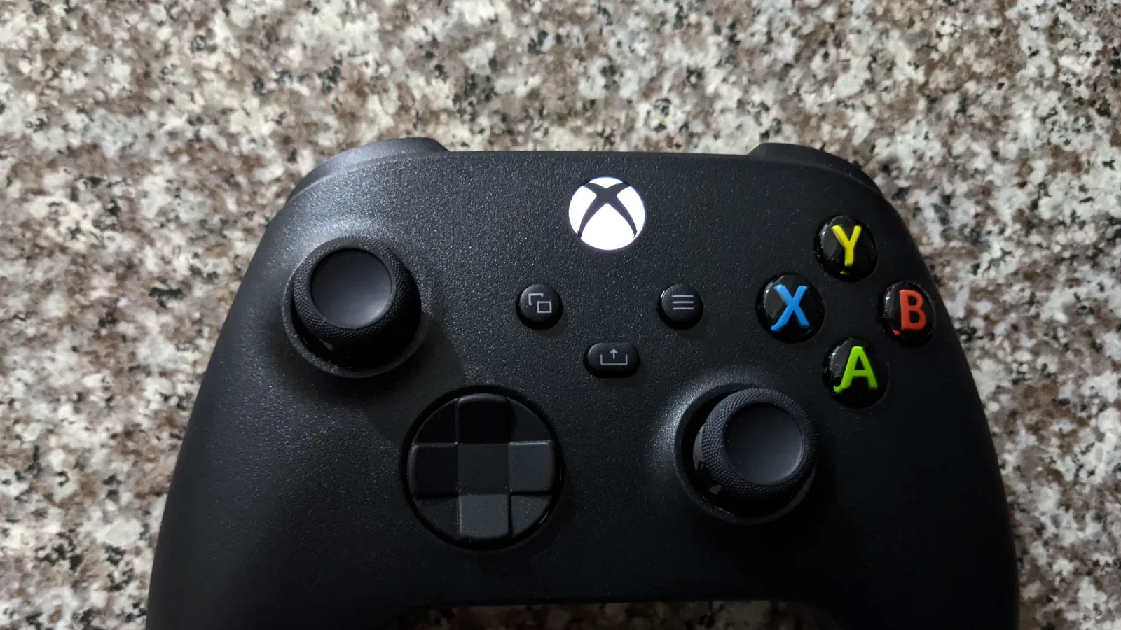 Xbox 무선 컨트롤러 전면부 클로즈업 사진 - 검은색 컨트롤러의 Xbox 버튼과 A·B·X·Y 버튼, 아날로그 스틱이 대리석 바닥 위에 놓인 모습