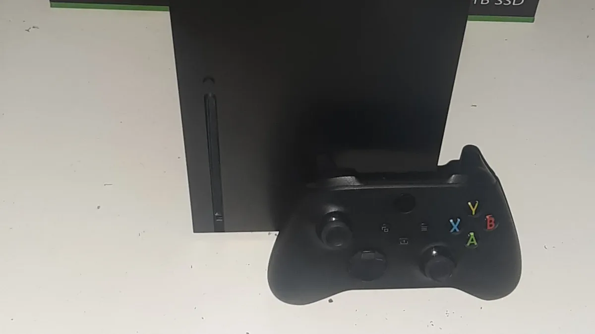 박스에서 막 꺼낸 검은색 Xbox Series X 본체와 컨트롤러가 나란히 놓인 사진 - 뒤로 Xbox 로고가 찍힌 포장 박스가 보인다