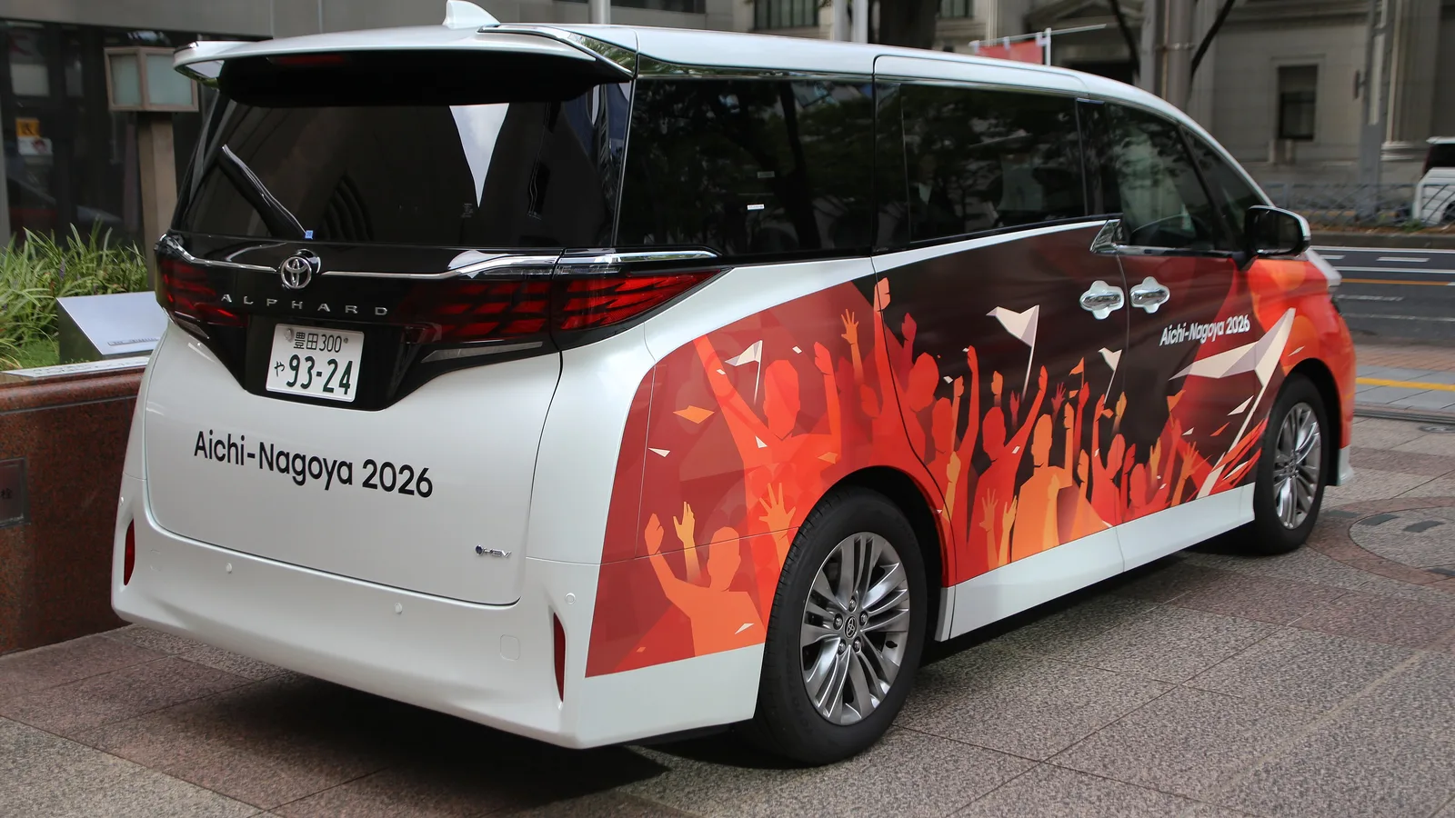‘Aichi-Nagoya 2026’ 문구와 붉은 관중 실루엣이 래핑된 도요타 알파드 승합차가 나고야 시내 보도에 서 있는 모습