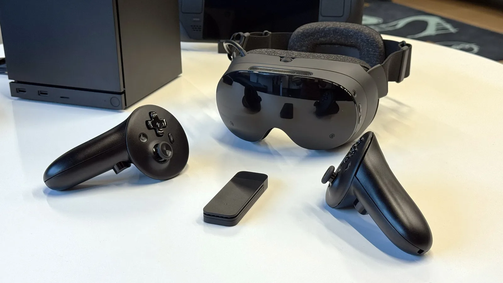 밸브 스팀 프레임 VR 헤드셋과 컨트롤러 실물 사진 - 흰색 스탠드얼론 헤드셋과 좌우 컨트롤러, 무선 동글이 테이블 위에 놓여 있는 모습
