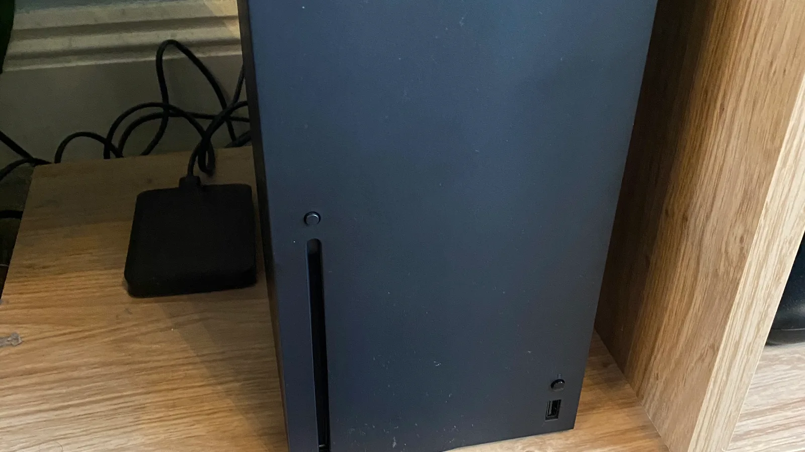 거실 선반 위에 세로로 세워진 검은색 Xbox 시리즈 X 콘솔과 그 옆에 놓인 Xbox 무선 컨트롤러